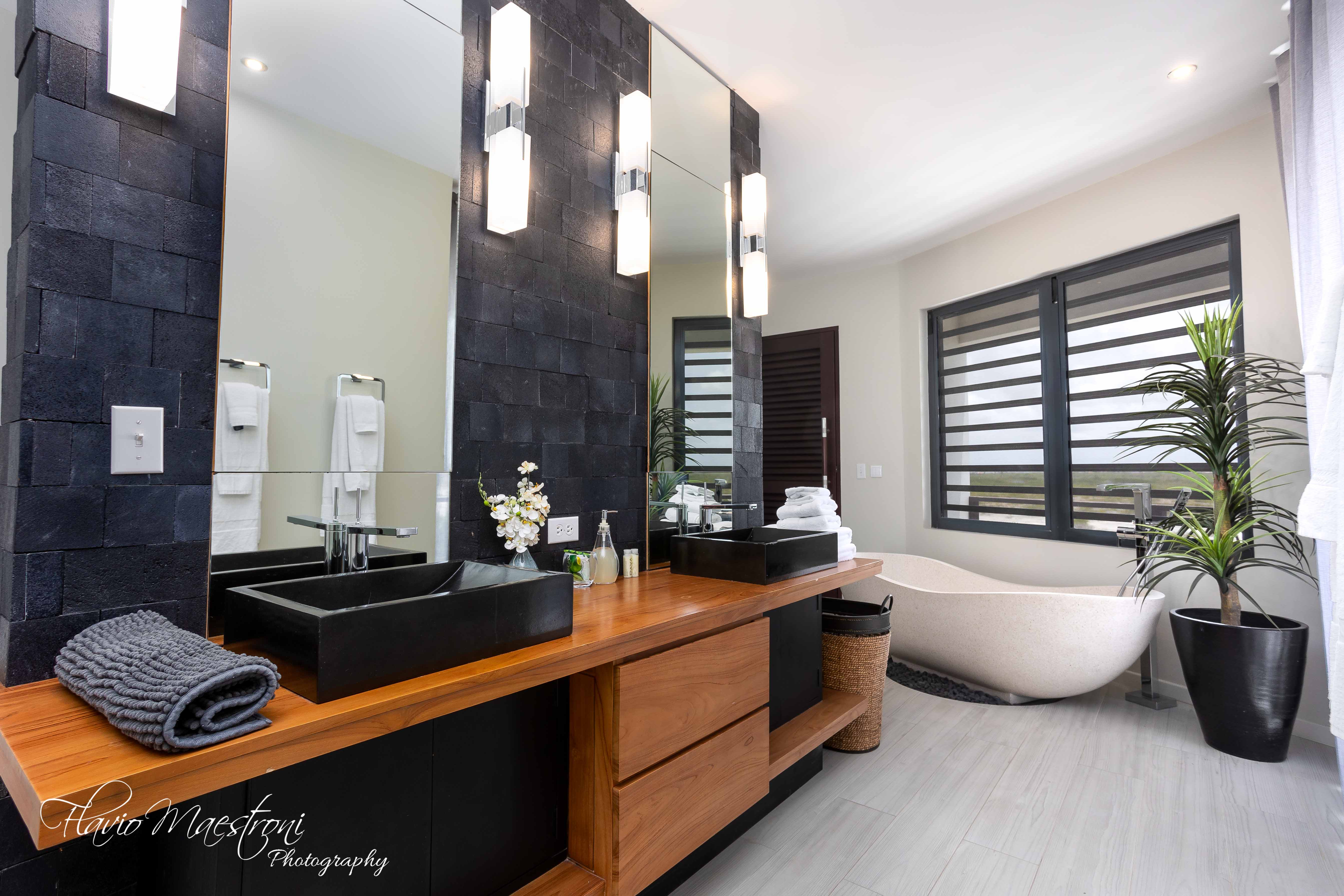 Luxury Bonaire villa spa bathroom with a freestanding soaking tub, double wood vanity with black vessel sinks, and a textured natural stone accent wall.